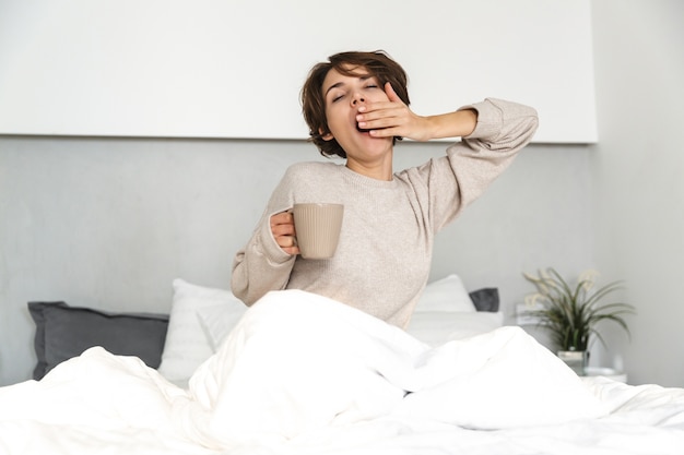 Lächelndes junges Mädchen, das sich morgens im Bett entspannt und Kaffeetasse hält