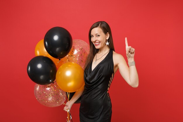 Lächelndes junges Mädchen im kleinen schwarzen Kleid, das den Zeigefinger feiert, der Luftballons auf rotem Hintergrund hält. Valentinstag Happy New Year Geburtstag Mockup Urlaub Party Konzept.