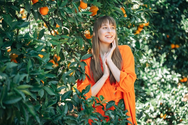 Lächelndes junges Mädchen in orangefarbenem Kleid schaut weg, indem es sich im orangefarbenen Garten an den Händen hält
