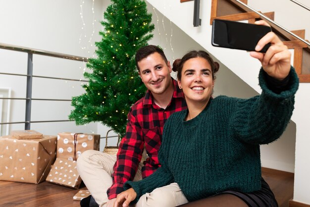 Lächelndes junges Paar macht Selfie mit Handy neben dem Weihnachtsbaum und präsentiert Holiday