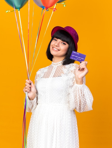Lächelndes junges Partygirl mit Partyhut, das Luftballons hält und Kreditkarte isoliert auf oranger Wand zeigt