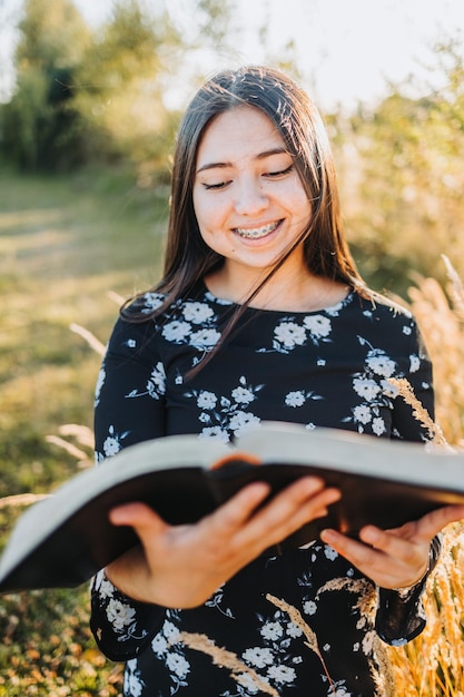 Lächelndes junges religiöses Mädchen, das ihre Bibel liest, draußen auf dem Feld bei Sonnenuntergang. Spirituelle Erweckung.