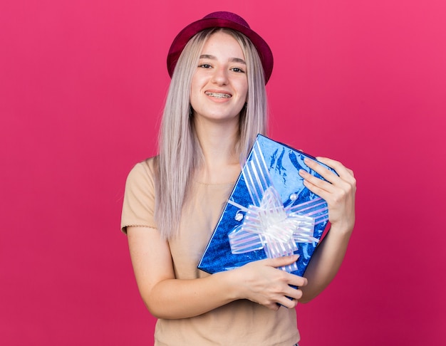 Lächelndes junges schönes Mädchen mit Partyhut mit Geschenkbox isoliert auf rosa Wand
