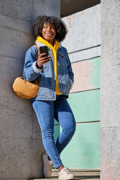 Lächelndes junges schwarzes Mädchen mit Afro-Haaren, das mit ihrem Smartphone in der Hand zur Seite schaut