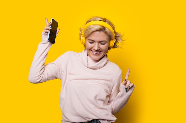 Lächelndes kaukasisches Mädchen mit blonden Haaren tanzt auf einem gelben Hintergrund, während Musik durch Kopfhörer hört