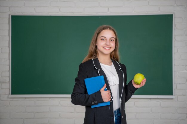 Lächelndes Kind, das in der Schule auf Tafelhintergrund mit Arbeitsbuch und Apfel steht