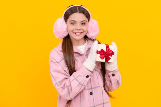 Lächelndes Kind in Ohrenschützern und Fäustlingen hält Geschenkbox auf gelbem Hintergrundkauf