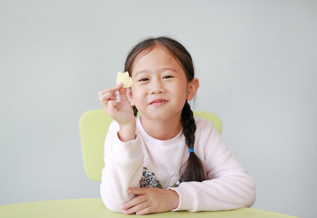 Lächelndes kleines asiatisches kindermädchen, das knusperige kartoffelchips auf weißem hintergrund isst. kind genießen, konzept zu essen.