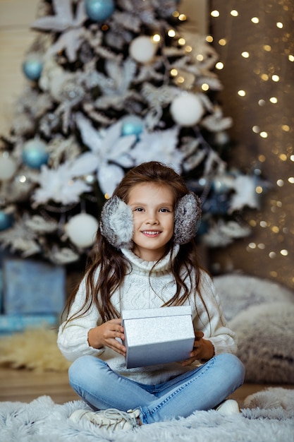 Lächelndes kleines Kindermädchen, das Geschenkbox nahe dekoriertem Weihnachtsbaum mit Lichtern hält
