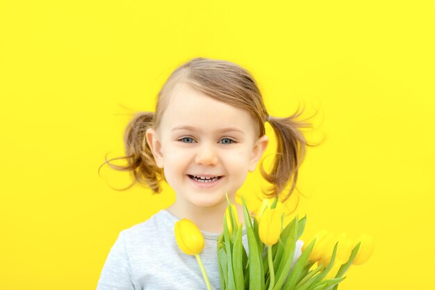 Foto lächelndes kleines kindermädchen im grauen kleid hält tulpen an einer gelben wand