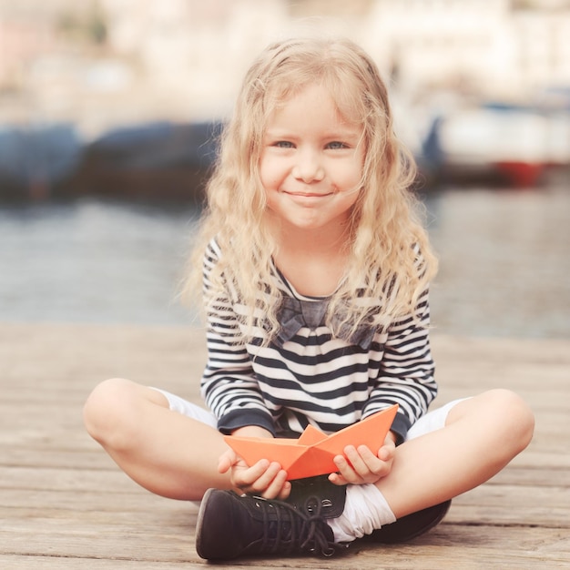 Lächelndes kleines Mädchen 5-6 Jahre alt, das draußen mit Papierboot spielt. Marineblaues Hemd tragen.