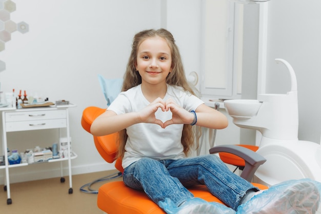 Lächelndes kleines Mädchen, das Herz mit den Händen im Zahnarztstuhl zeigt