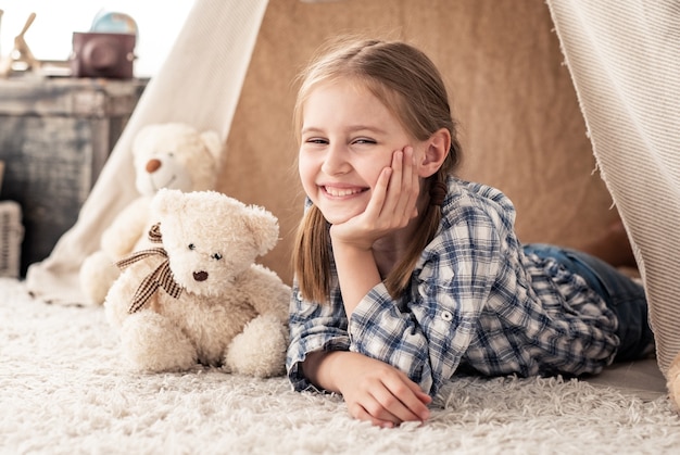 Lächelndes kleines mädchen, das im wigwam mit teddybären im kinderzimmer liegt