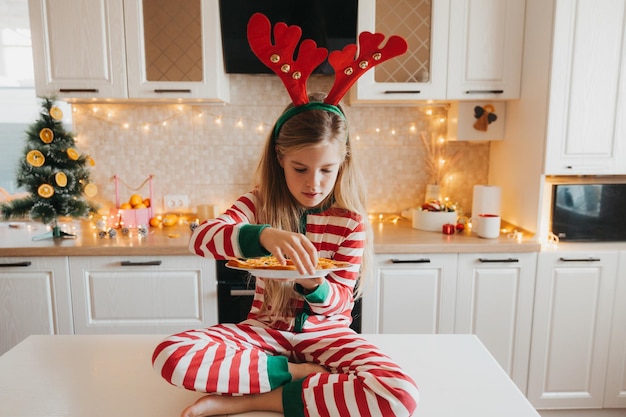 Lächelndes kleines Mädchen, das mit Orangen am weißen Tisch in der Küche sitzt. Kinderspiele in der dekorierten Küche zu Weihnachten. Glückliches kleines Mädchen an Heiligabend zu Hause