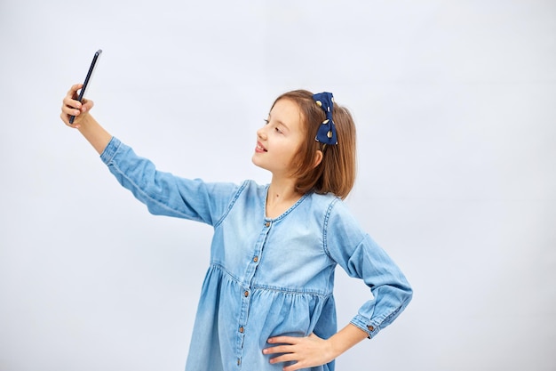 Lächelndes kleines Mädchen im lässigen Jeanskleid hält Smartphone und macht Selfie