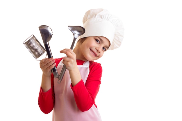 Lächelndes kleines Mädchen im roten Hemd mit weißer Schürze und Hut, die Küchengeräte auf weißem Hintergrund halten
