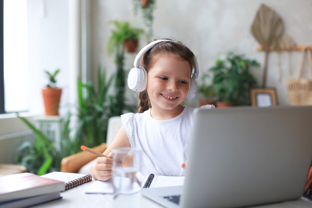 Lächelndes kleines Mädchen in Kopfhörern handschriftliche Studie online mit Laptop zu Hause, süßes glückliches kleines Kind in Kopfhörern nimmt Internet-Web-Unterricht oder -Klasse auf dem PC.