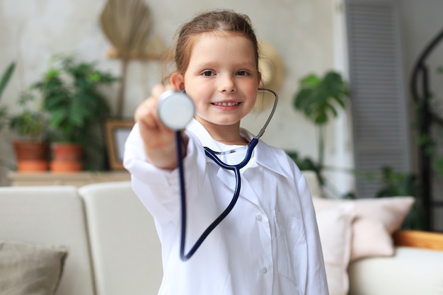 Lächelndes kleines Mädchen in medizinischer Uniform, das zu Hause mit Stethoskop spielt.