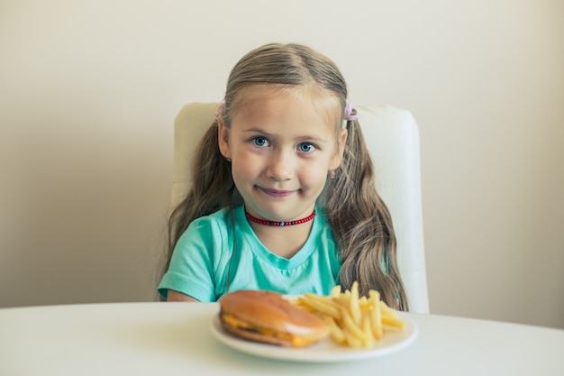 Lächelndes kleines Mädchen sitzt am Tisch mit Hamburger und Pommes-Frites-Gericht