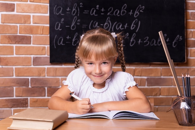 Lächelndes kleines Studentenmädchen, das an einer Schulbank sitzt und Mathe studiert