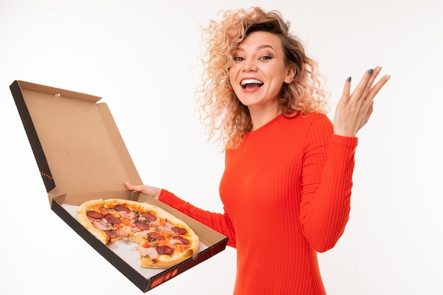 Lächelndes lockiges blondes Mädchen in einem roten Kleid hält eine Schachtel Pizza auf weißer Wand