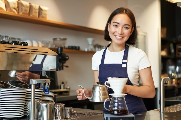 Lächelndes Mädchen Barista asiatischer Barkeeper gießt Wasser aus Wasserkocher Filterkaffee im Café hinter der Theke mit blauer Schürze