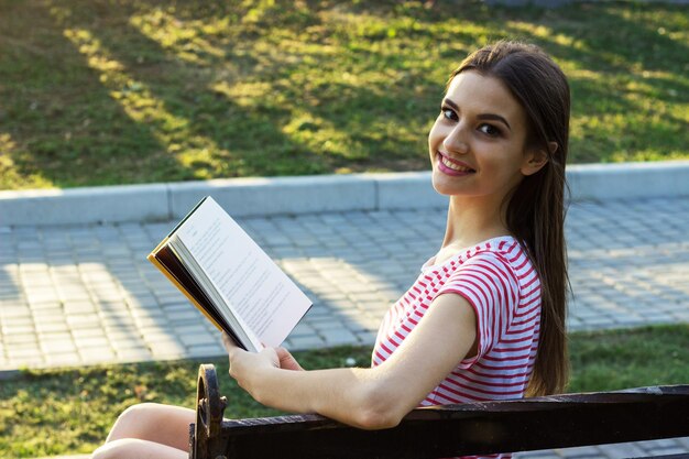 Lächelndes Mädchen, das auf einer Holzbank sitzt und ein Buch liest Hübsches junges Mädchen dreht den Kopf beim Lesen eines Buches im Park nach hinten Seitenansicht