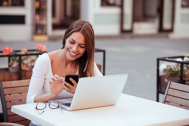 Lächelndes Mädchen, das draußen Telefon und Laptop im Café verwendet.