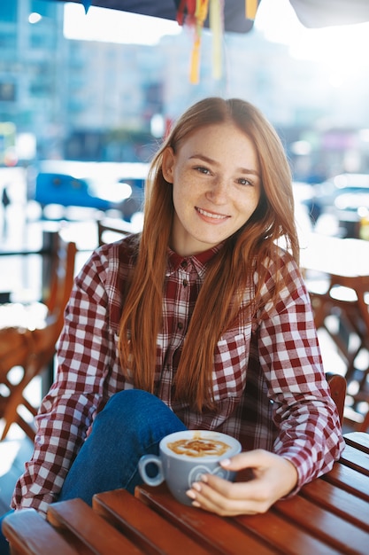 Lächelndes Mädchen, das eine große Tasse Kaffee hält, während auf Kamera schaut