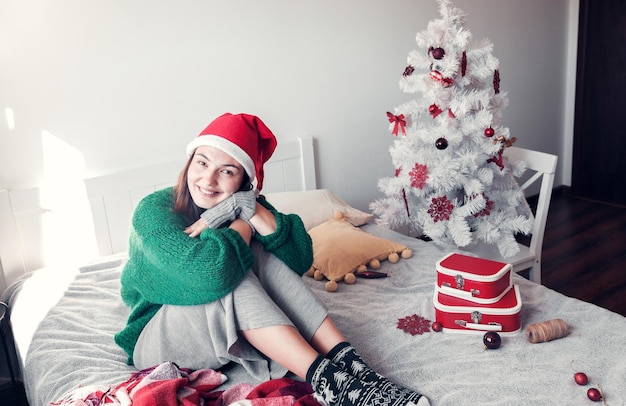 Lächelndes Mädchen, das einen Weihnachtsmann-Hut trägt, sitzt auf einem Bett vor dem Hintergrund eines Weihnachtsbaums