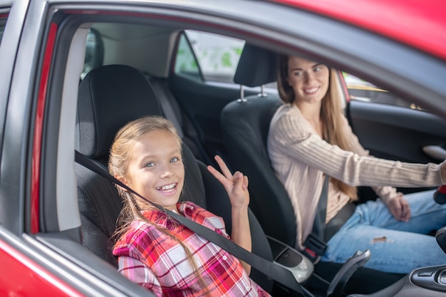 Lächelndes Mädchen, das Friedenszeichen auf Beifahrersitz zeigt und mit ihrer Mutter mit dem Auto fährt