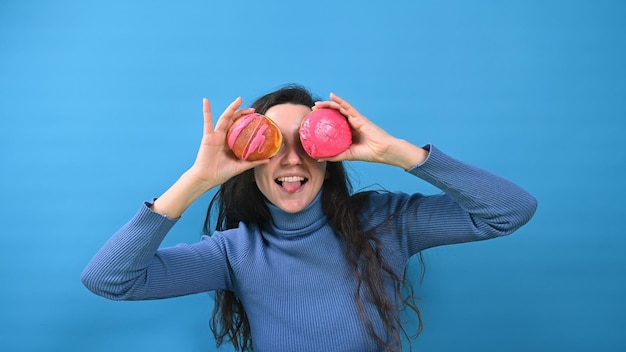 Lächelndes Mädchen, das seine Augen mit verglasten Donuts bedeckt Hübsche Frau, die Spaß auf blauem Hintergrund mit Kopierraum hat