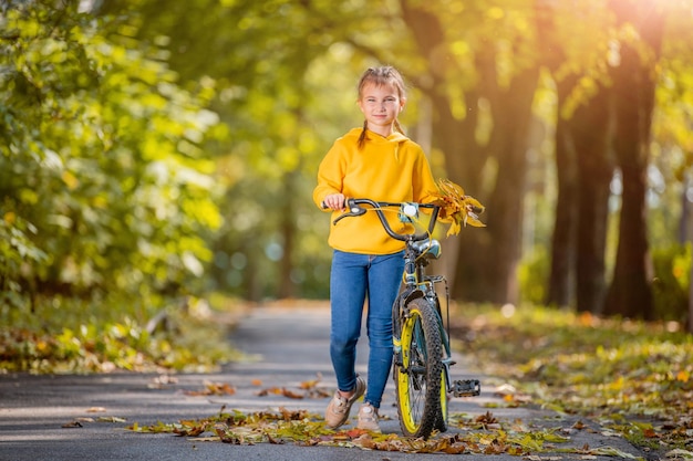 Lächelndes Mädchen im gelben Pullover geht an einem sonnigen Tag mit dem Fahrrad in einem Herbstpark spazieren und winkt freundlich mit der Hand