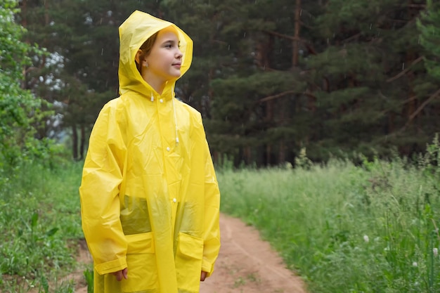 Lächelndes Mädchen im Regenmantel steht auf der Straße gegen grünen Wald