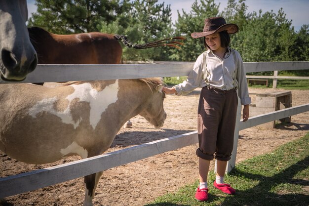 Lächelndes Mädchen in der Cowboykleidung streichelt kleines Pony im Fahrerlager am sonnigen Sommertag. Kümmert sich um Tiere Konzept.