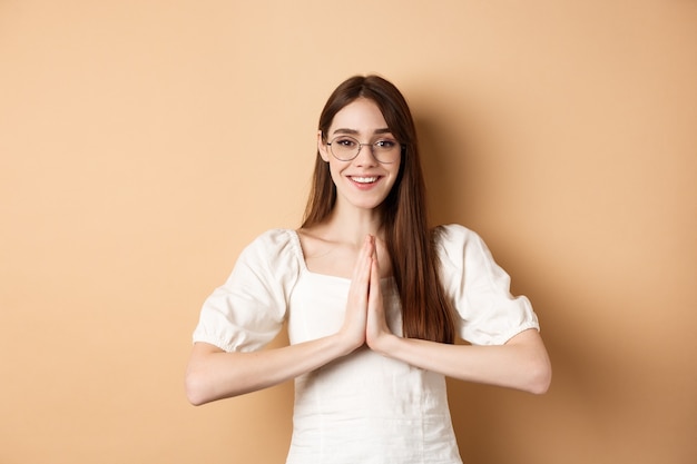 Lächelndes Mädchen in Gläsern sagen Danke, zeigen namaste Geste, drücken Dankbarkeit aus, stehen erfreut auf beigem Hintergrund.