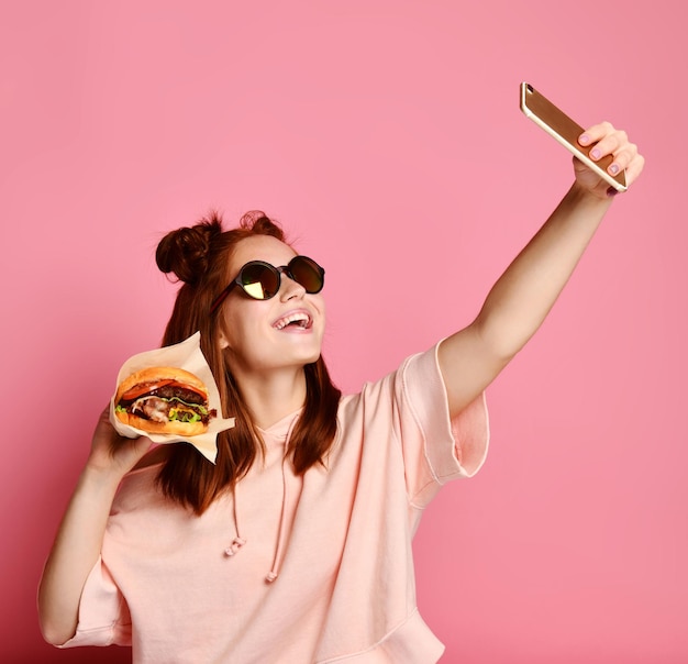 Lächelndes Mädchen in Lifestyle-Kleidung, das ein Selbstporträt auf dem Smartphone mit Burger-Sandwich macht