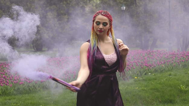 Lächelndes Mädchen in lila Satinkleid mit langen bunten Zöpfen und auffälligem Glitzer-Make-up. Purpurroter Rauch bedeckt das Mädchen im Frühlingspark