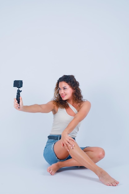Lächelndes Mädchen macht Selfie mit ihrer Minikamera, indem sie auf dem Boden auf weißem Hintergrund sitzt