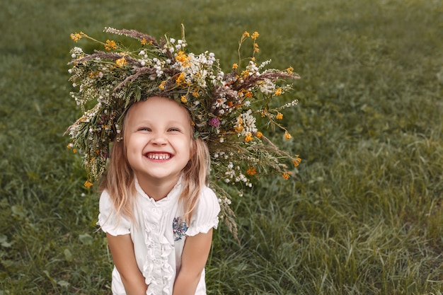 Lächelndes Mädchen mit einem Blumenkranz auf dem Kopf auf einem Grashintergrund schaut auf cmera