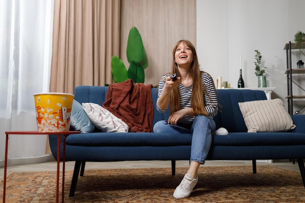 Lächelndes Mädchen mit Fernbedienung in der Hand, das auf dem Sofa fernsieht