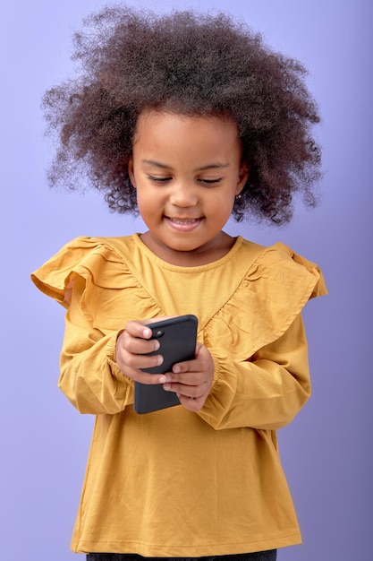 Lächelndes Mädchen mit lockigem Haar, das mit dem Smartphone auf den Bildschirm des Mobiltelefons schaut, isoliert auf lila
