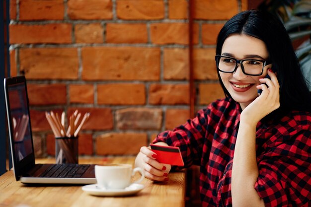 Lächelndes Mädchen mit schwarzen Haaren, die Brillen tragen, die im Café mit Laptop, Handy, Kreditkarte und Tasse Kaffee sitzen, freiberufliches Konzept, Online-Shopping, rotes Hemd tragend.