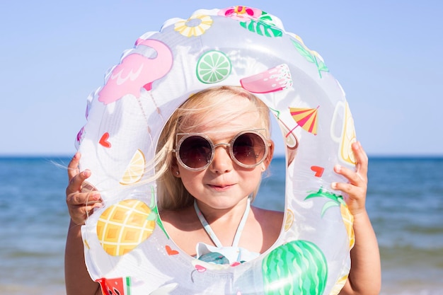 Lächelndes Mädchen mit Sonnenbrille sieht durch einen aufblasbaren Kreis am Strand