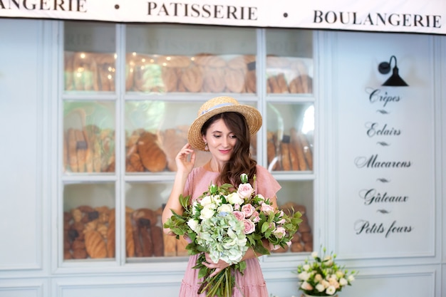 lächelndes Mädchen steht in der Nähe der Fensterbäckerei in Strohhut mit einem Blumenstrauß Überraschungsgeschenk