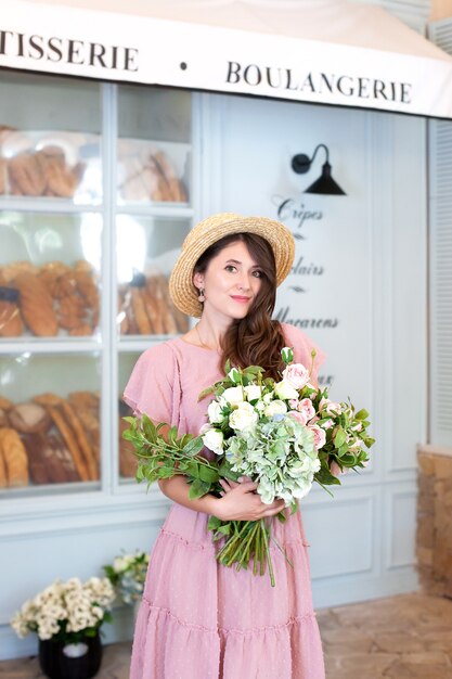 lächelndes Mädchen steht in der Nähe der Fensterbäckerei in Strohhut mit einem Blumenstrauß Geschenk