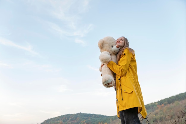 Lächelndes Mädchen umarmt Teddybär geht durch bergiges Gelände Junge Frau im gelben Regenmantel mit weichem Spielzeug draußen