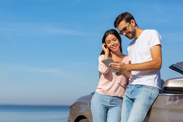 Lächelndes Mädchen und Kerl, die im Smartphone schauen, während sie das Auto an einem sonnigen Tag lehnen