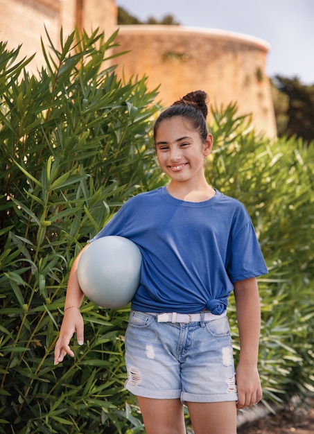 Lächelndes Mädchenkind, das T-Shirt und Jeansshorts trägt, bleibt mit einem Ball im Freien im Park