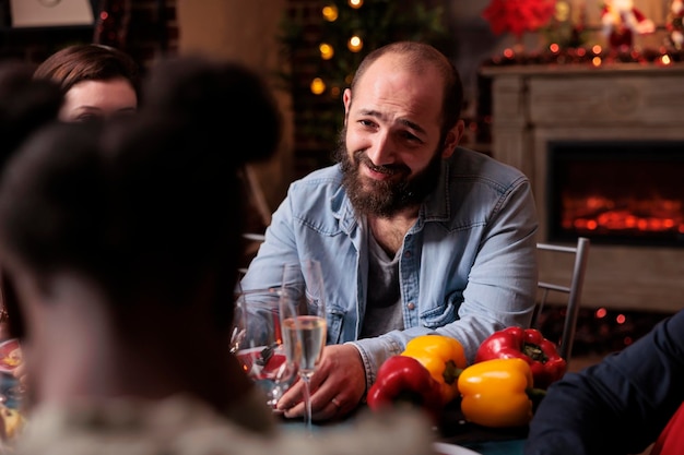 Lächelndes Mannporträt beim festlichen Weihnachtsessen, Winterurlaub mit der Familie zu Hause feiernd, Kamin im Hintergrund. Person, die am Tisch sitzt, Sekt trinkt, Weihnachtsessen isst